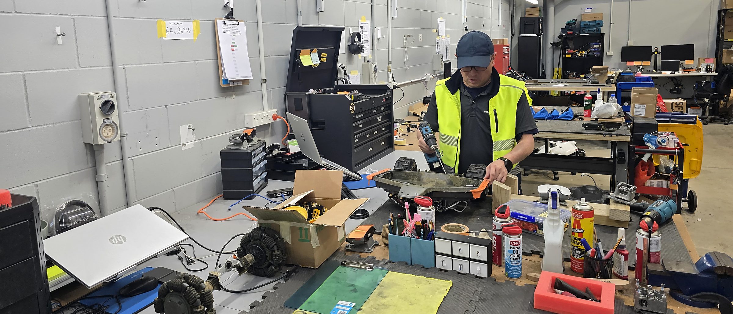 A Silk Lane Group technician at the bench with a power drill, working on a stripped robotic mower chassis, with parts and tools laid out around him